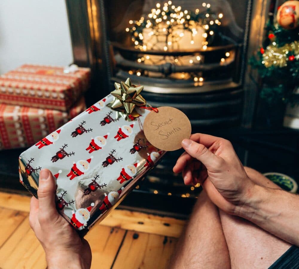 holding a gift with a secret santa tag near the fireplace and with more gifts on the side