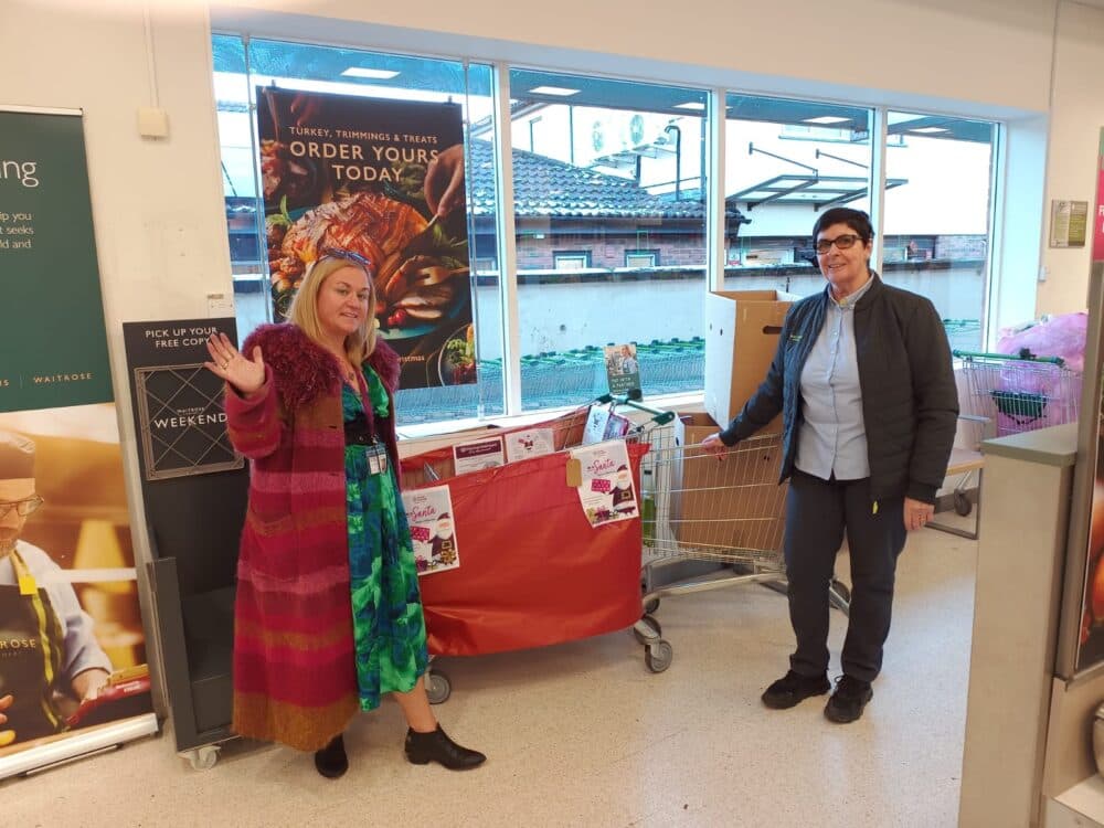 Two women stand by a shopping cart with boxes and flyers inside a grocery store, smiling at the camera. - Home Instead
