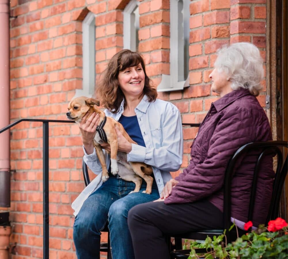 A senior woman sitting outside while talking with with her daughter who had emdium lenght hair and holding a dog both happy