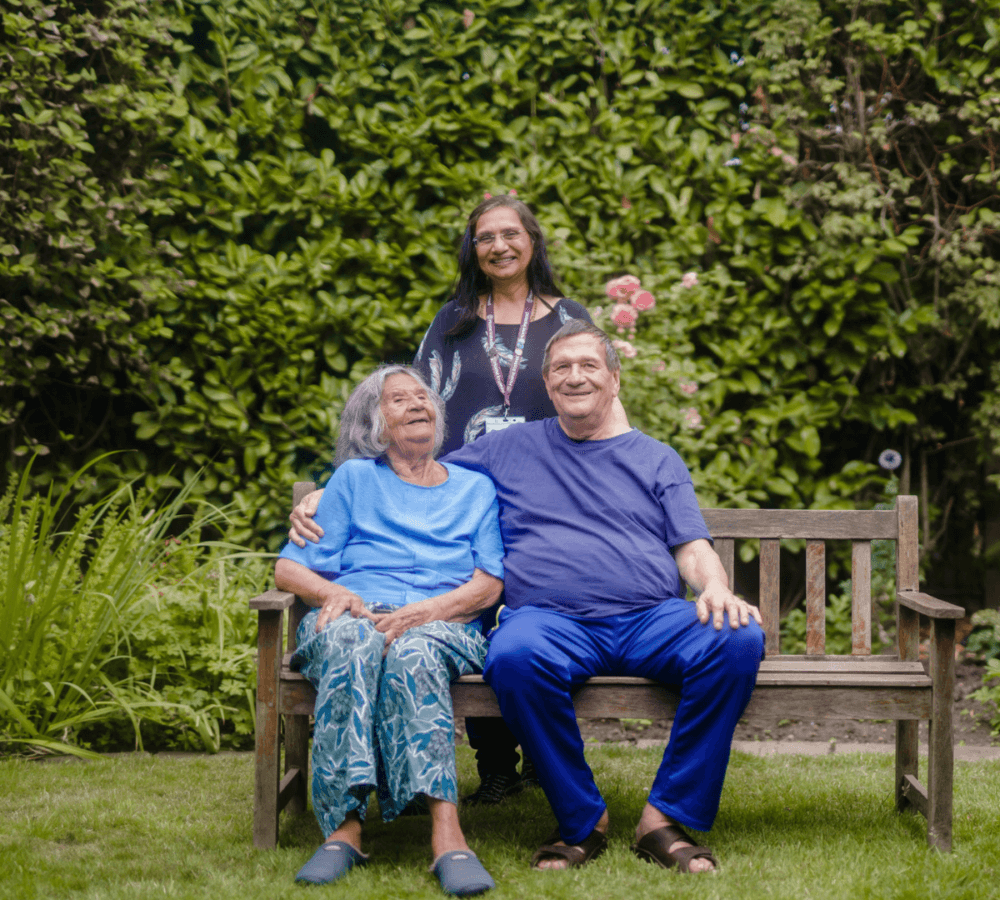 Senior mum and son sitting on a bench with carer standing at the back outside the garden with green surroundings