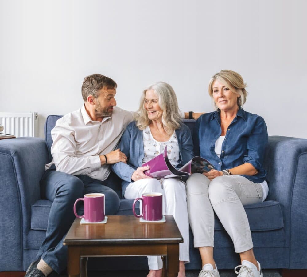 Mum and son looking at each other both smiling and sitting on the couch with her daughter beside her and holding a home care brochure with Home Instead cups on the table