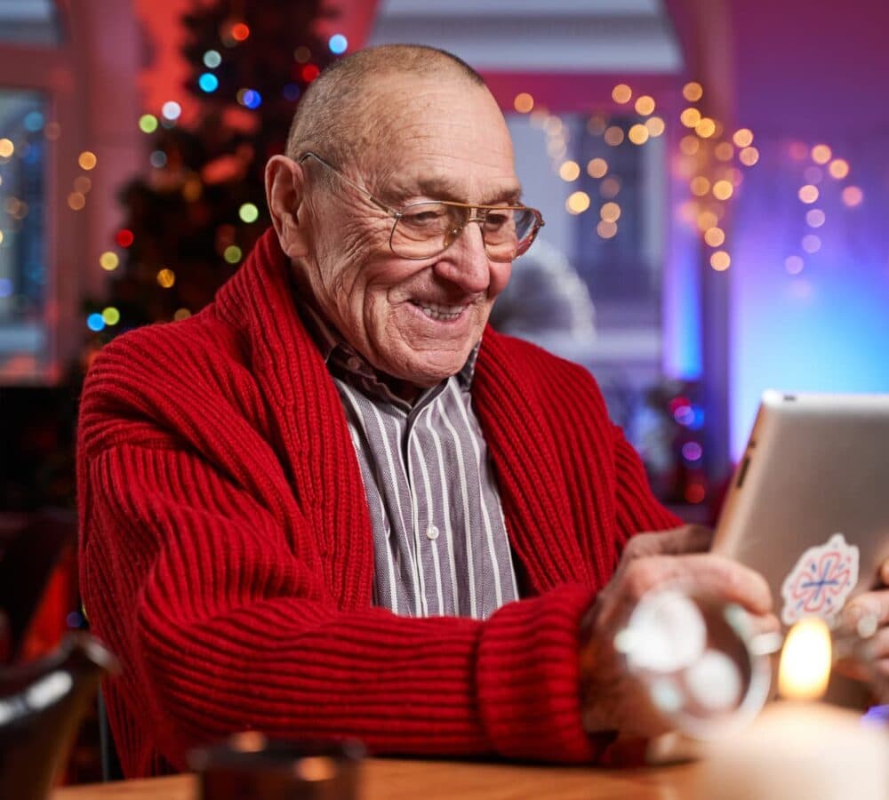 Senior man happy holding a gadget on the table with Christmas tree and lights on the background