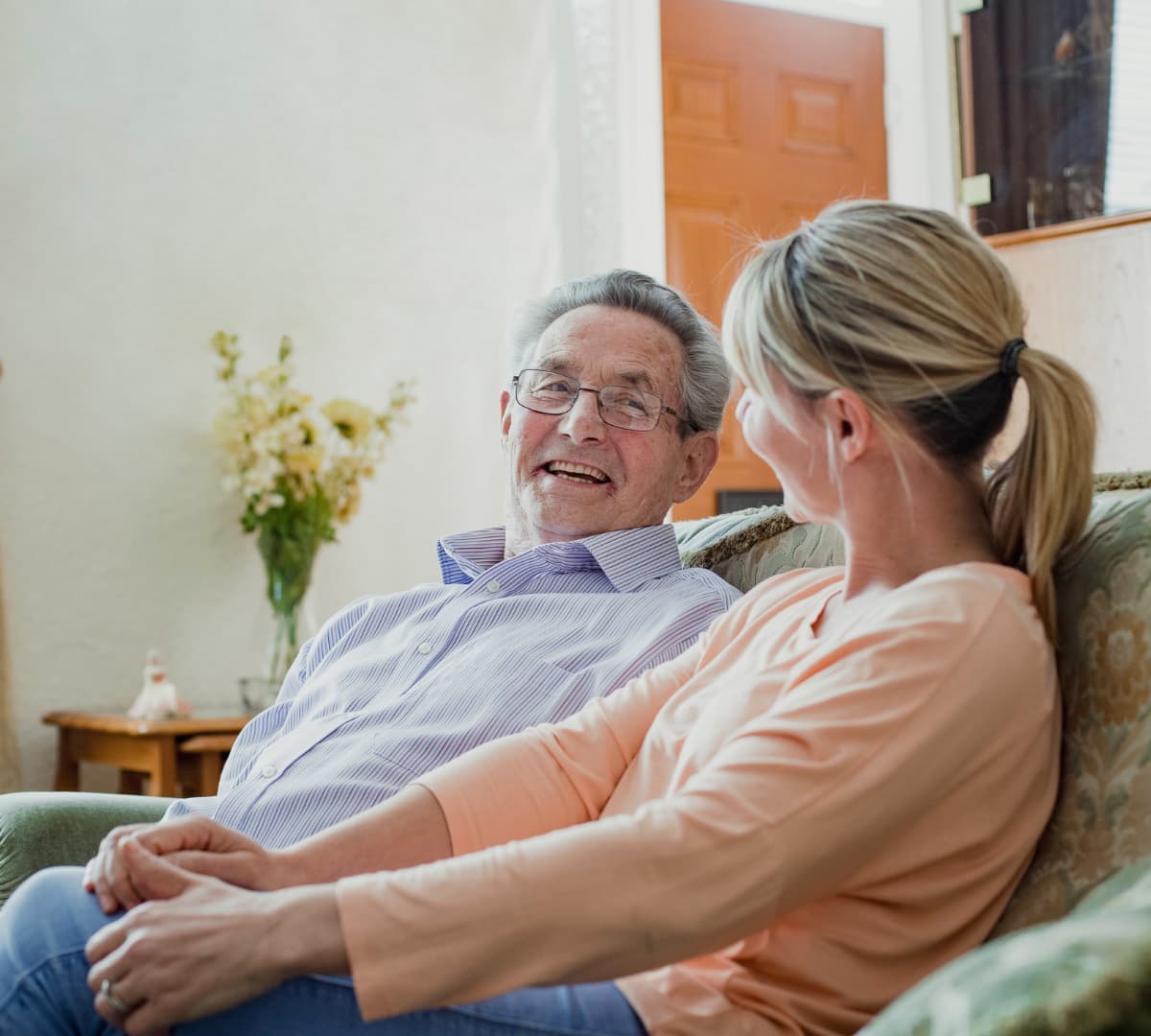 Senior man wearing eyeglasses sitting on a couch with his daughter beside him and having a conversation inside the house with a flower vase at the background