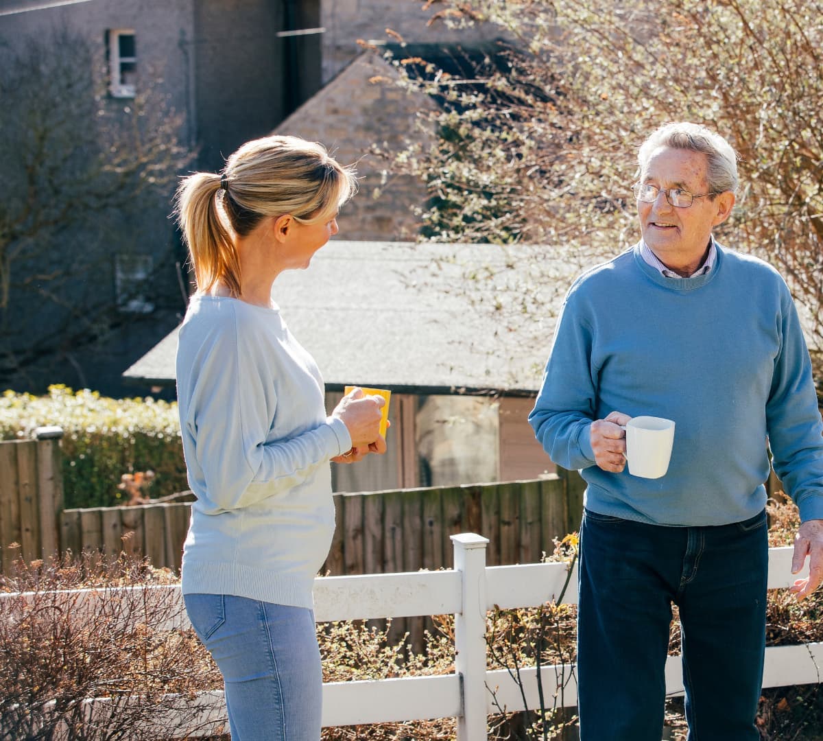 A senior man with daughter outside the house enjoying fresh air and both holding a cup of coffee