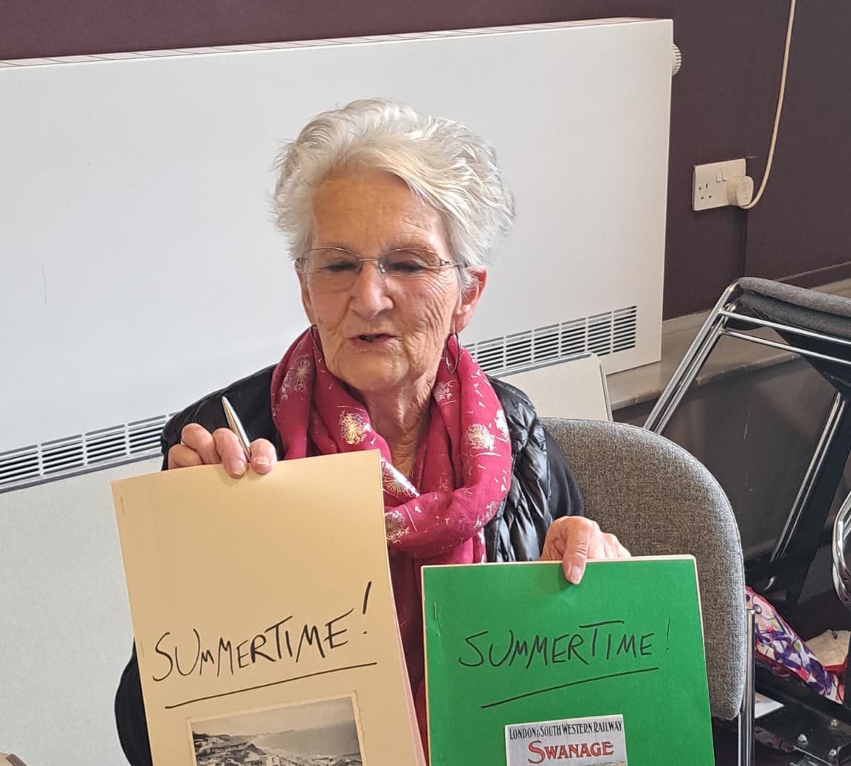 Elderly woman wearing eyeglasses and a scarf holding her two poem while sitting down
