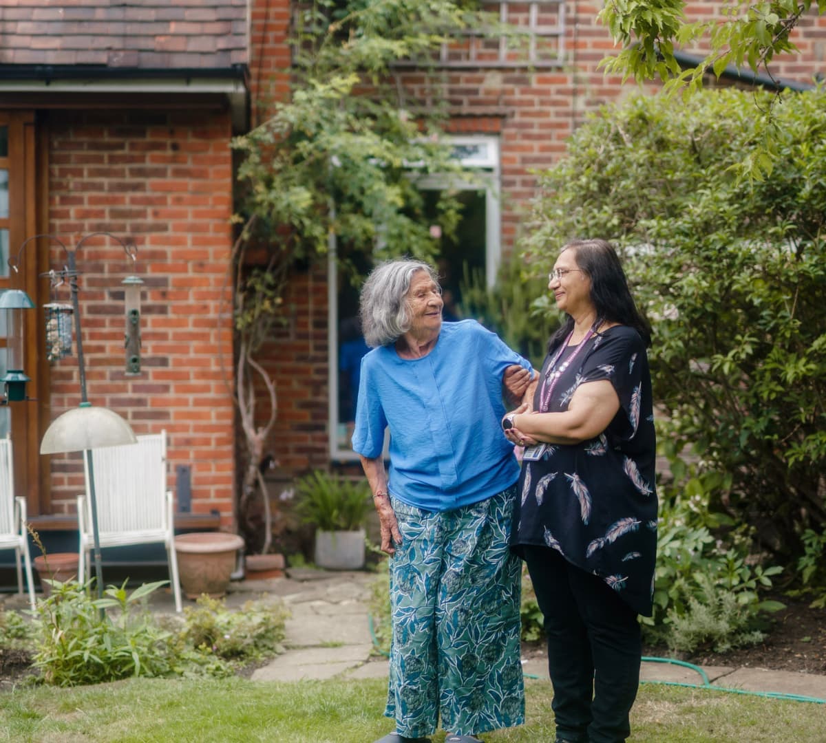 Senior woman in the garden wearing blue shirt walking with her carer wearing black and with long hair