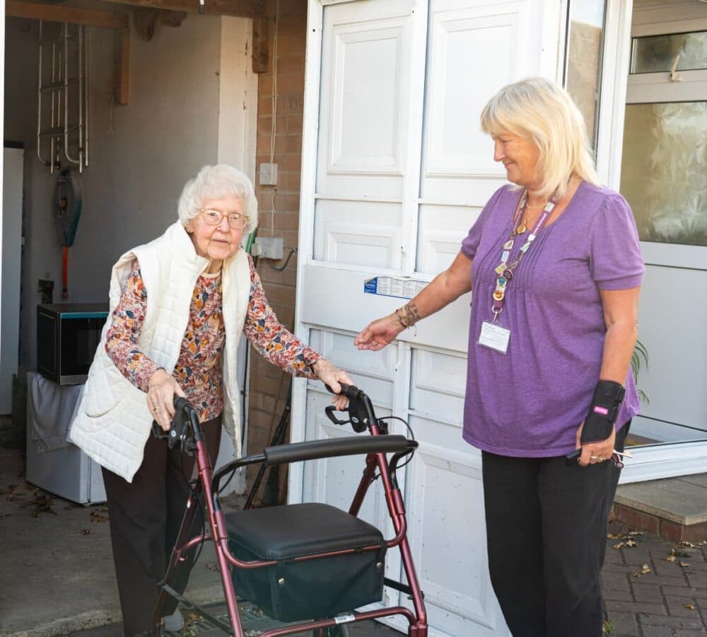 senior woman with short hair and eyeglasses going out of the door using a walker with her carer wearing purple