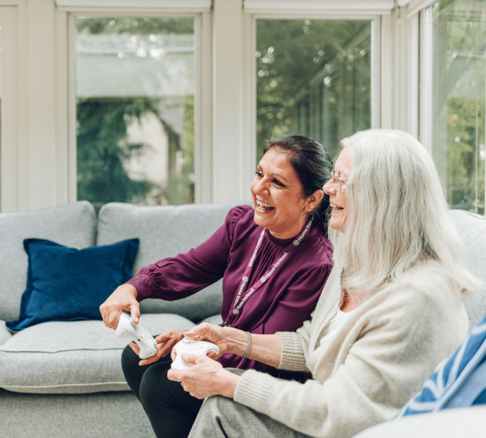 Senior woman with carer both happy while playing video games inside the house while sitting on the couch with some glass windows in the background