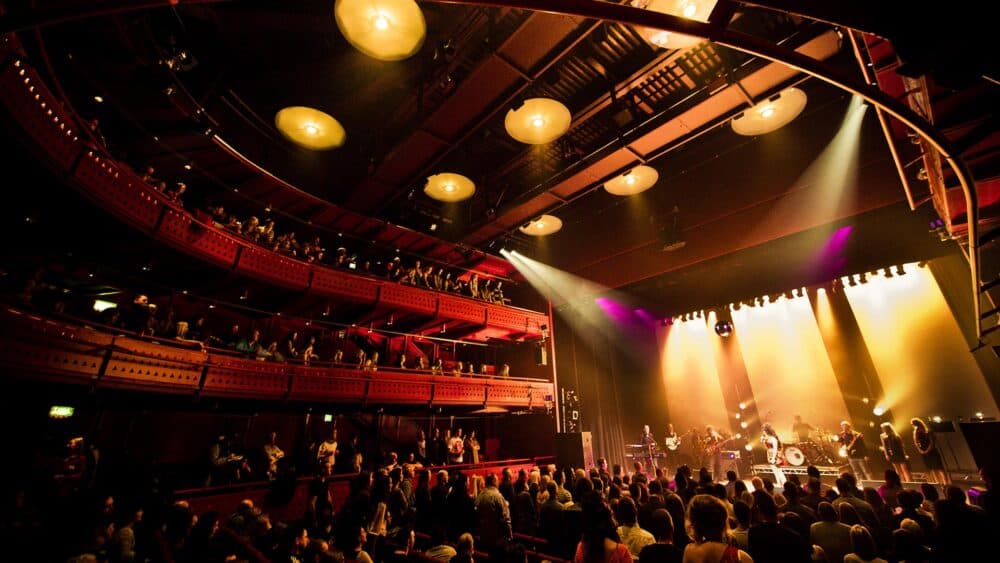 A lively audience watches a band perform on stage in a theater with balcony seating and warm lighting. - Home Instead