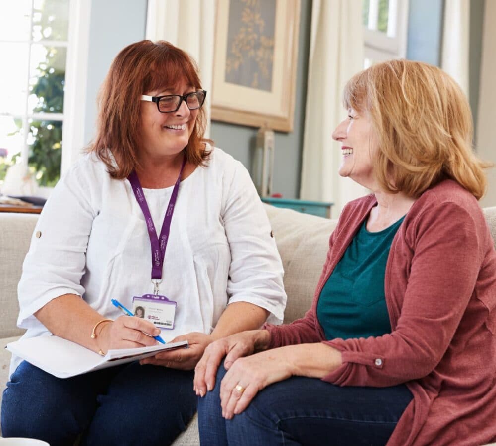 Two women sitting on a couch while talking and smiling. The other woman wearing eyeglasses and holding a notebook