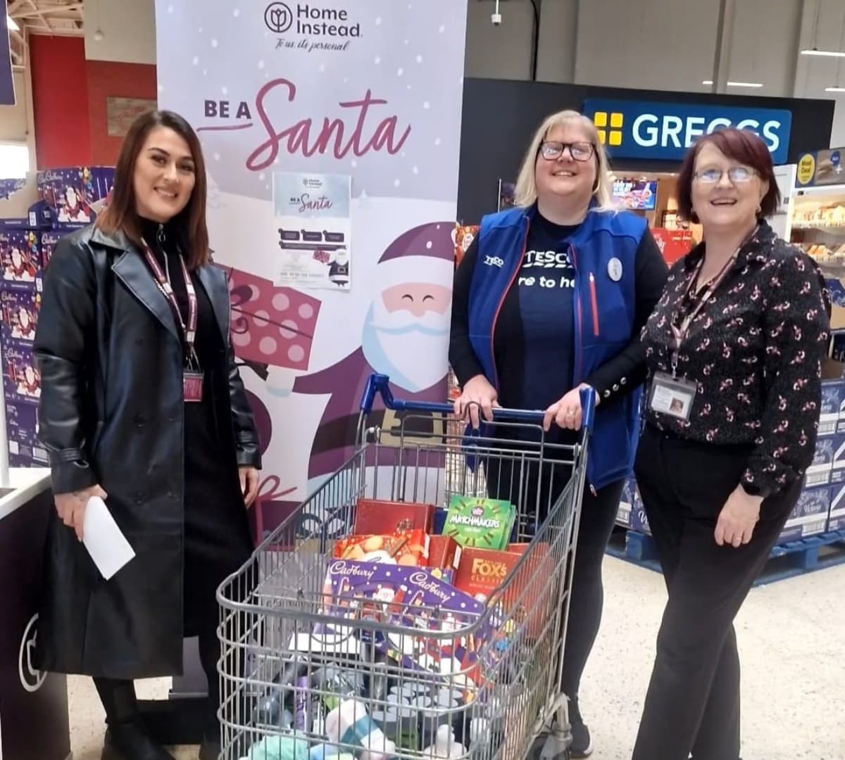 Three ladies with a Be a Santa banner at the background and holding a shopping cart