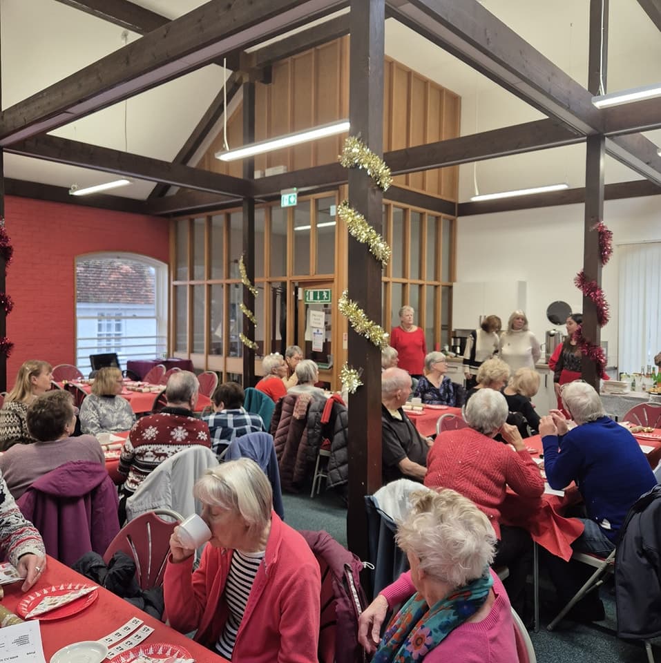 A group of older adults gather in a decorated hall, sitting at tables and chatting during a festive event. - Home Instead