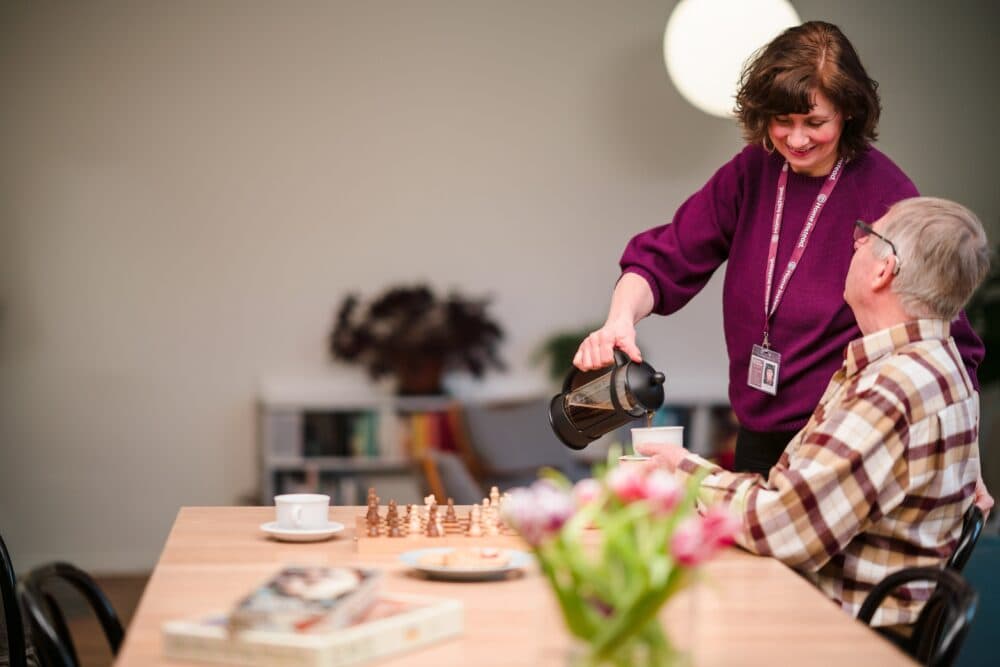 A woman pours coffee for an older man sitting at a table with books, flowers, and a chessboard. - Home Instead