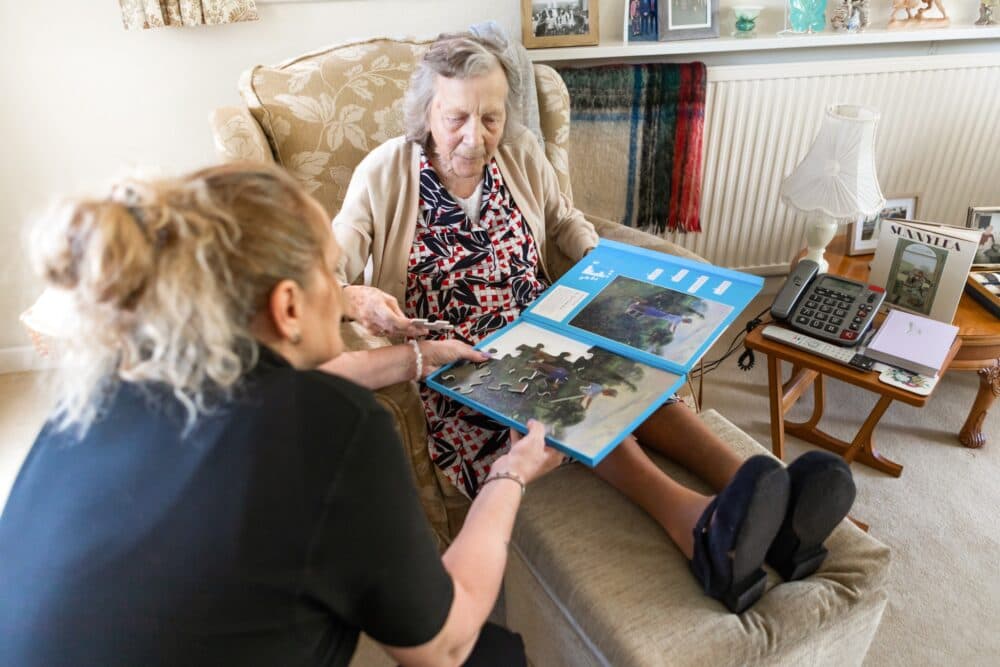 An elderly woman and a caregiver work on a jigsaw puzzle together in a cozy living room. - Home Instead