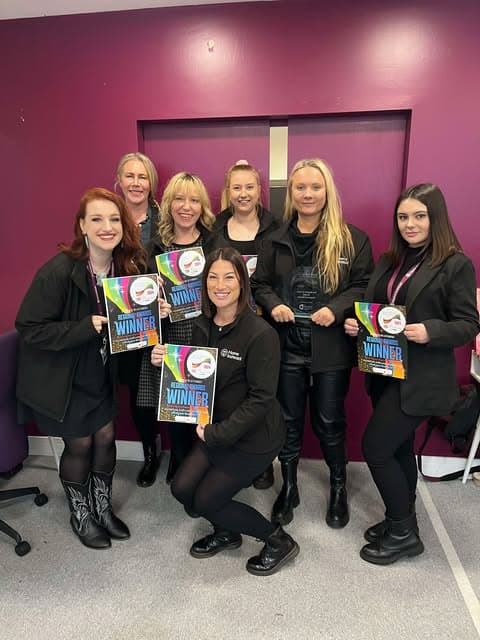Six women smiling and holding "Winner" certificates, standing in front of a purple wall. - Home Instead