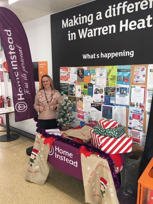 Woman stands at a Home Instead table with gifts and decorations, in front of a bulletin board. - Home Instead