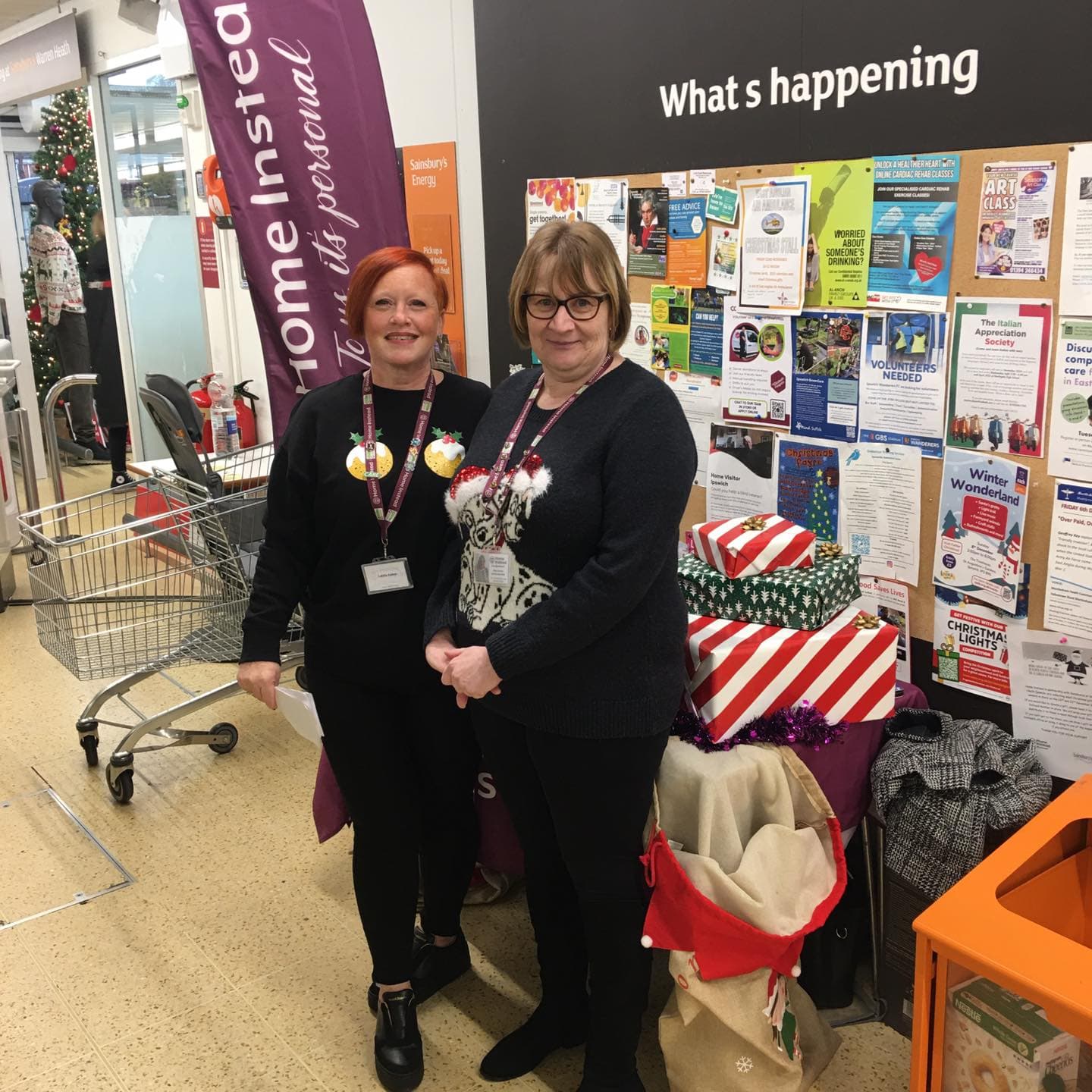 Two women stand by a festive donation table with gift boxes and a community notice board behind them. - Home Instead