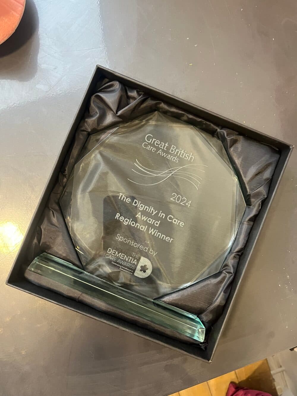 A glass award in a box reads "Great British Care Awards 2024, The Dignity in Care Award, Regional Winner. - Home Instead