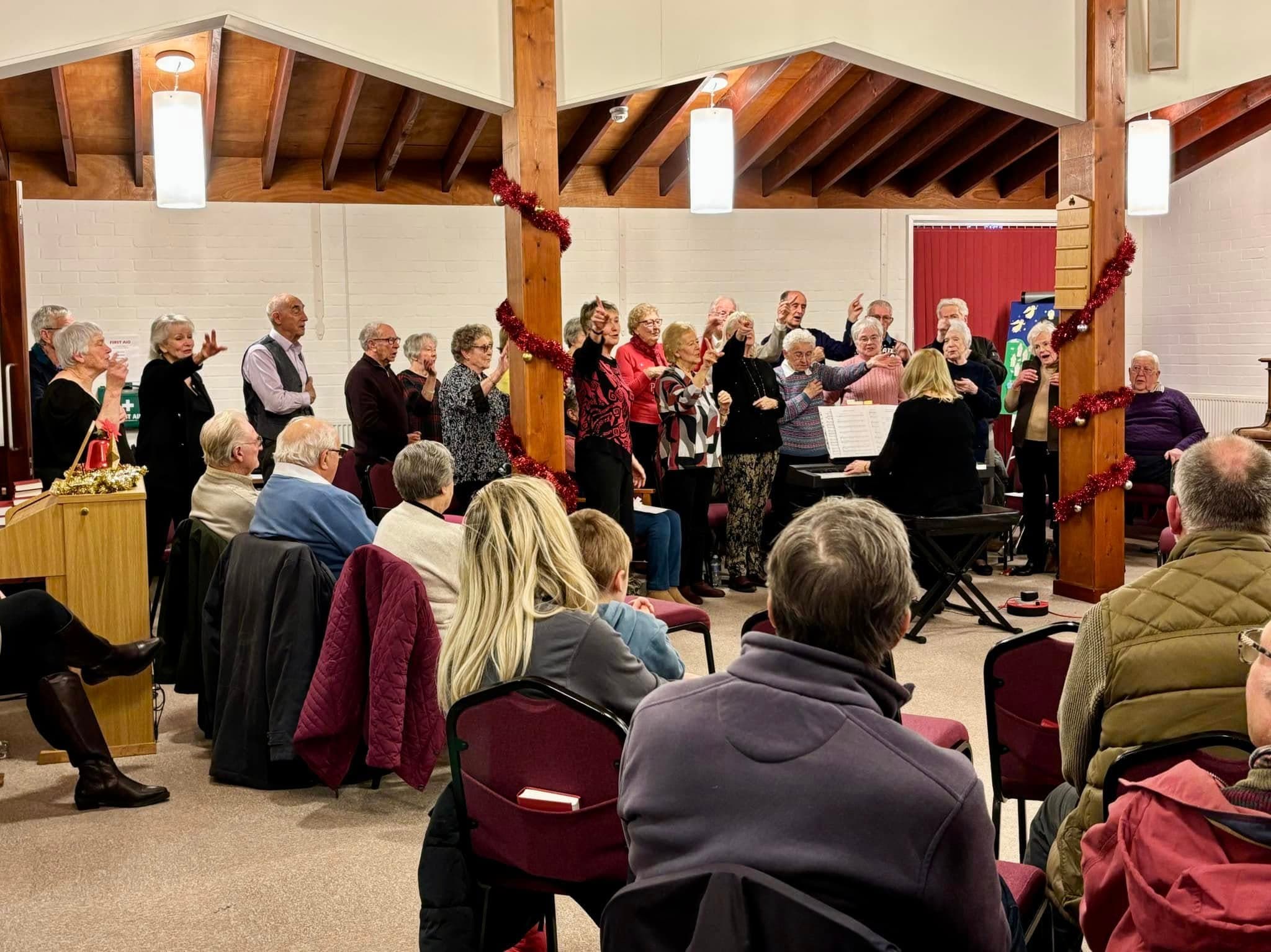 A choir performs in a church hall while an audience watches from red chairs. - Home Instead