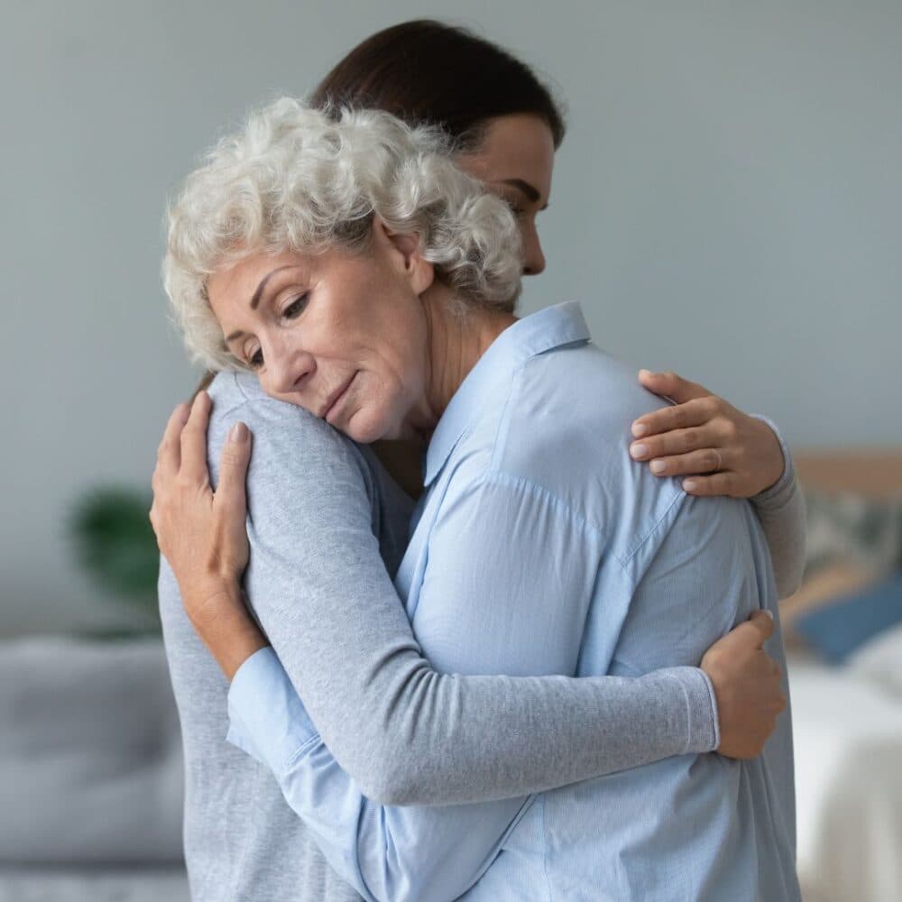 Elderly woman and younger woman hugging each other closely, both appearing emotional and comforted. - Home Instead