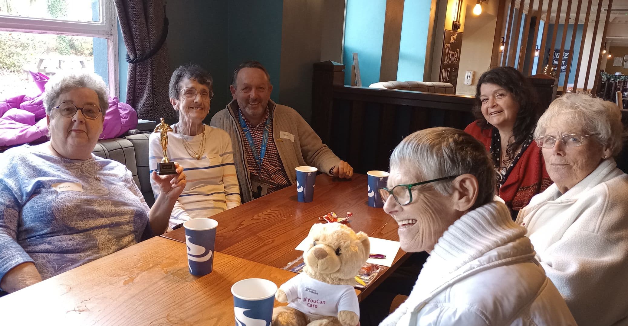 Six smiling older adults sit around a table with drinks and a teddy bear, one holding a trophy. - Home Instead