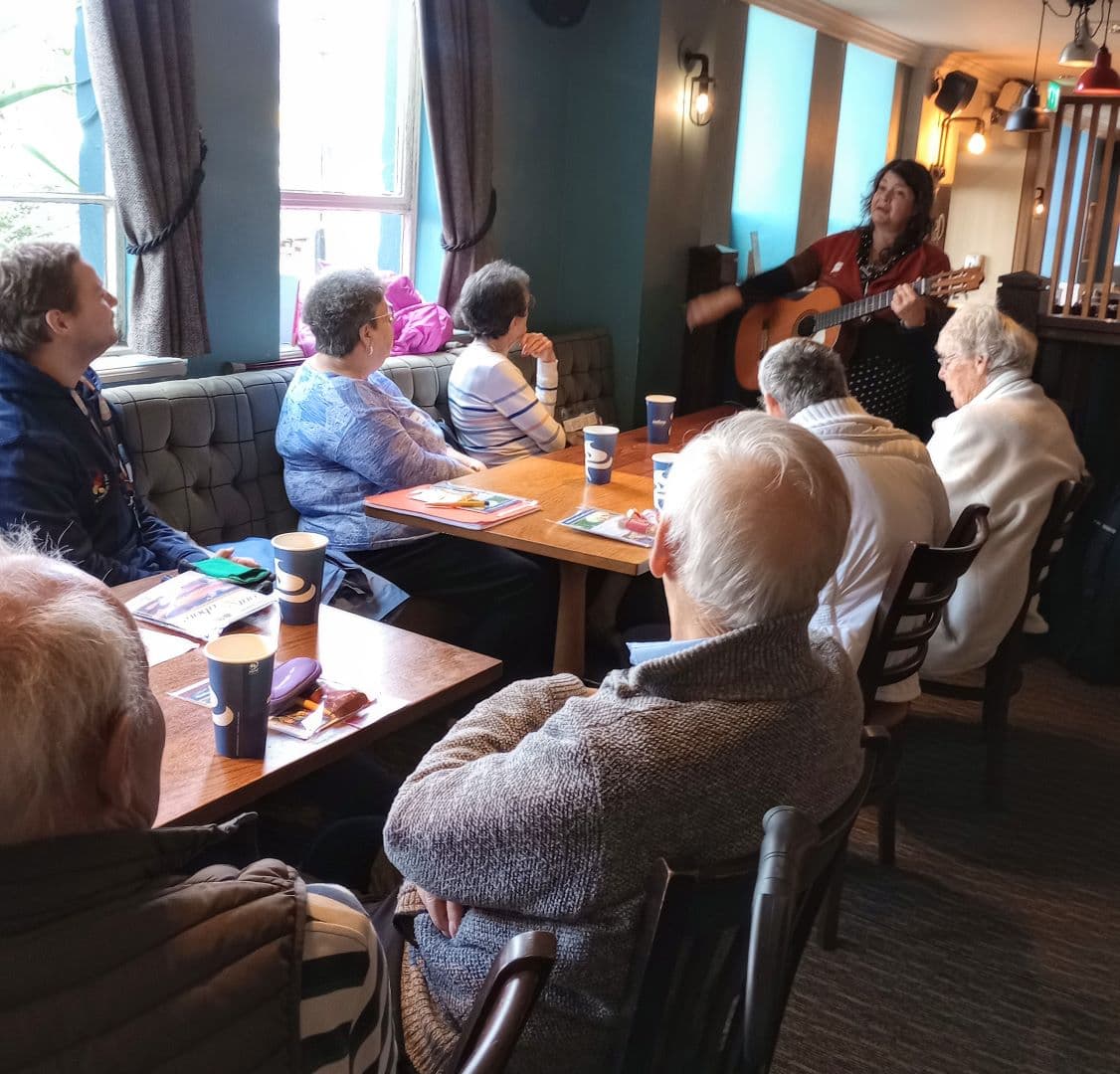 A woman plays guitar and sings to a group of elderly people seated around tables in a cozy room. - Home Instead