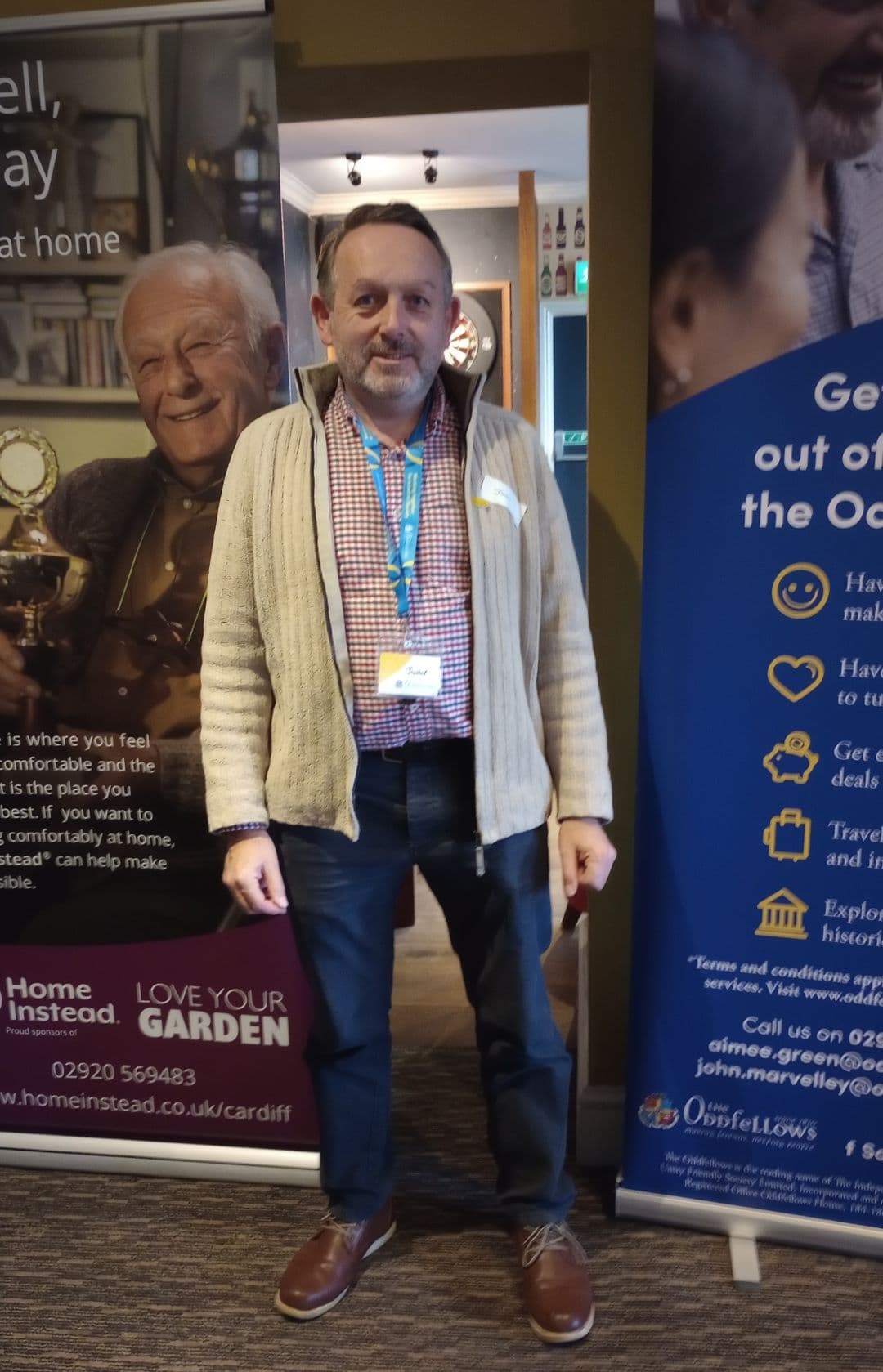 A man stands indoors between two promotional banners, wearing a beige sweater, blue lanyard, and brown shoes. - Home Instead