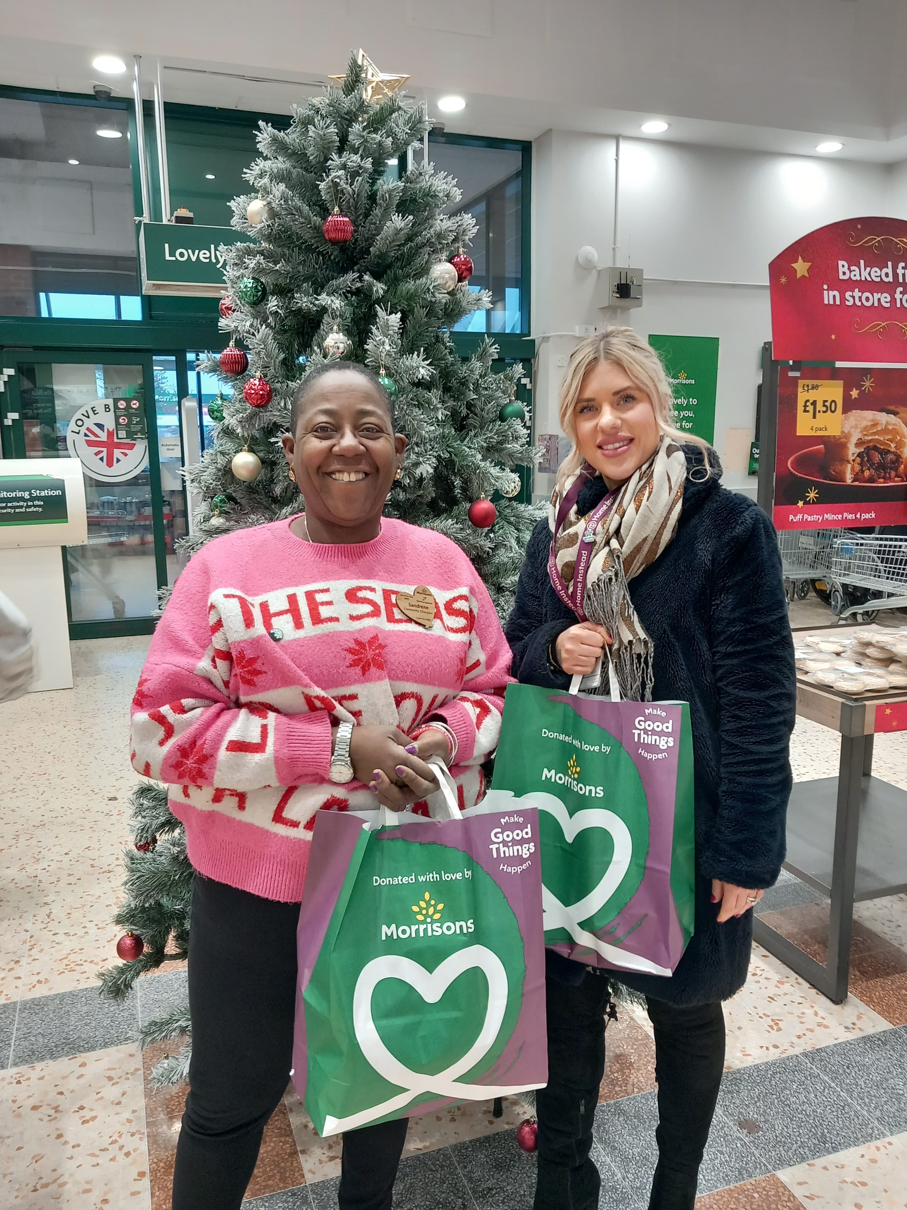 Two women smiling, holding Morrisons shopping bags, stand by a Christmas tree inside a supermarket. - Home Instead