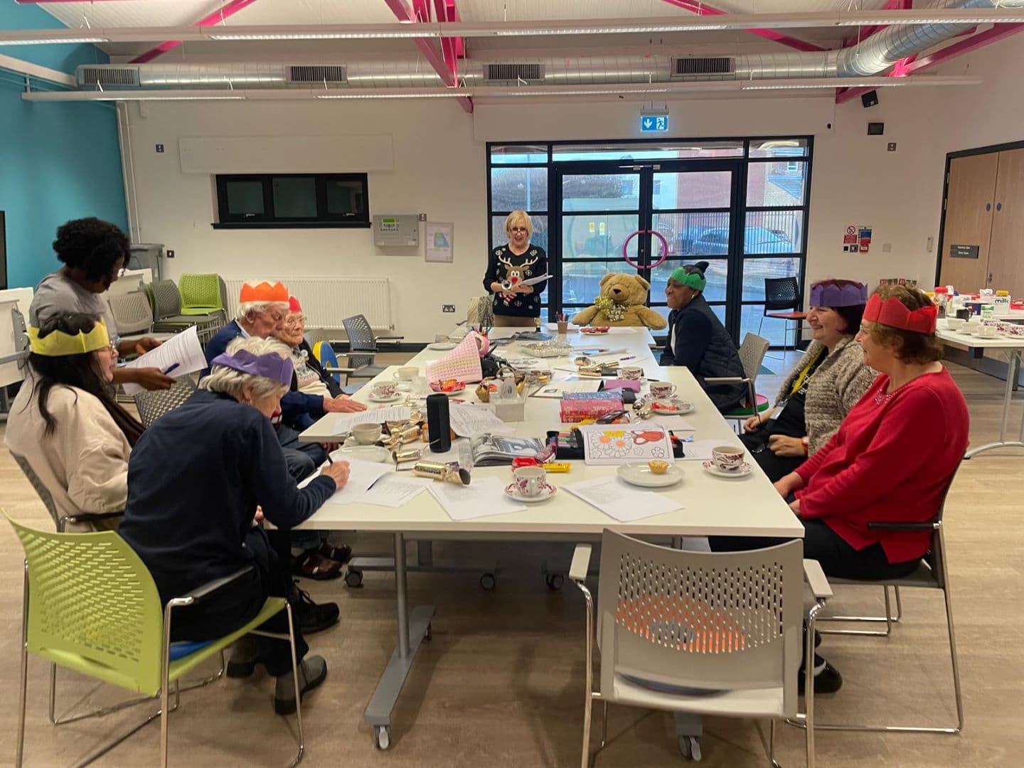 A group of people wearing festive hats sit around tables in a bright room, engaged in an activity. - Home Instead