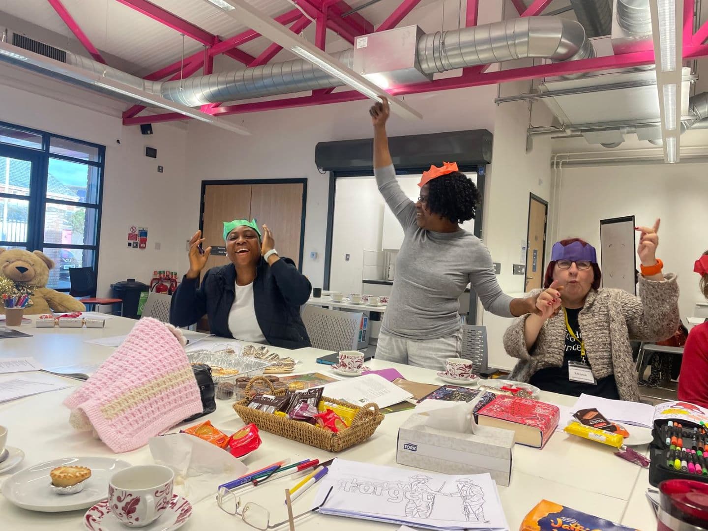 Three women at a table laugh and dance wearing colorful paper hats, surrounded by snacks and craft supplies. - Home Instead