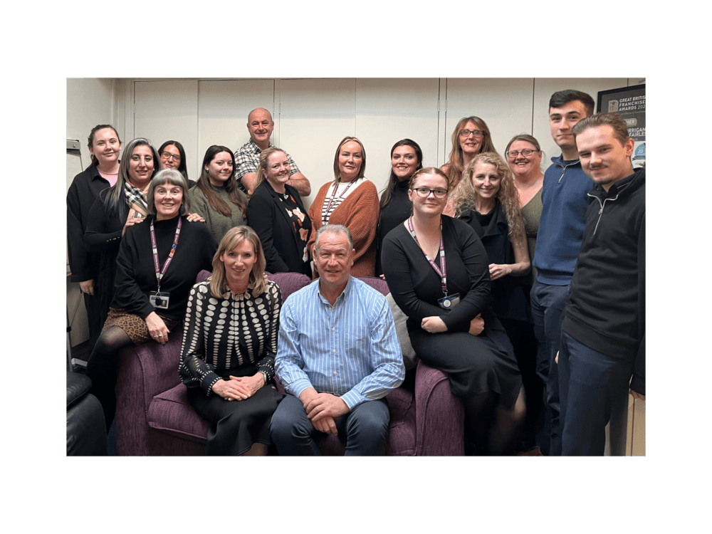 A group of people, some seated and some standing, pose together indoors and smile at the camera. - Home Instead