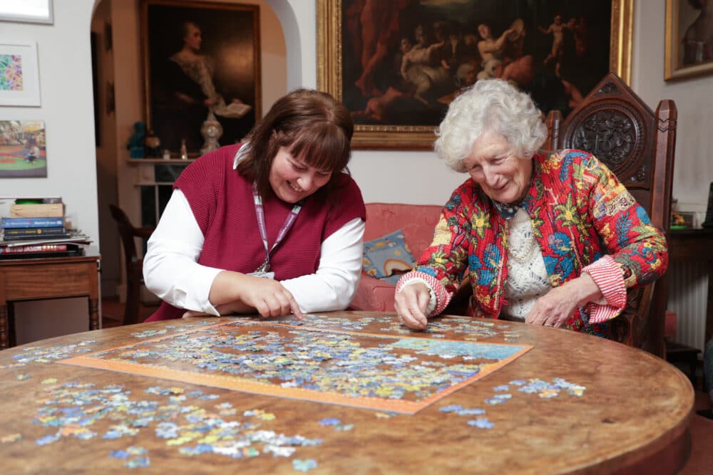Two women, one elderly, smiling and working on a jigsaw puzzle together at a round wooden table. - Home Instead