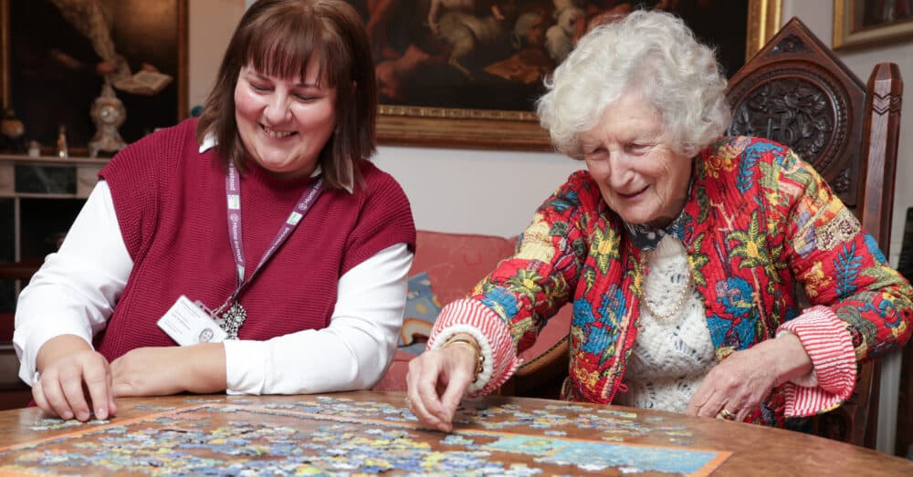 Two women smiling and working together on a jigsaw puzzle at a table in a cozy room. - Home Instead