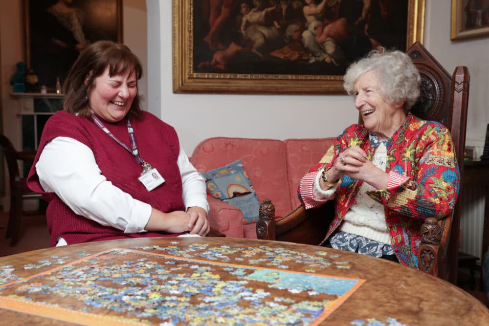 Two women smiling and laughing together while working on a jigsaw puzzle at a table in a cozy room. - Home Instead