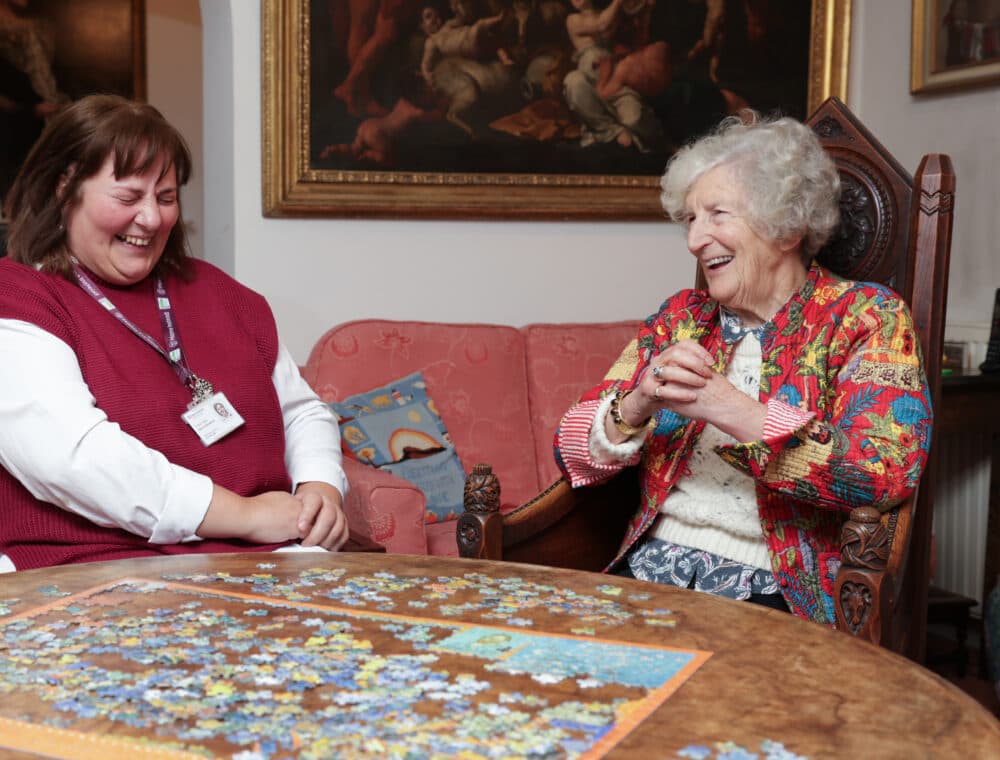 Two women smile and laugh together while working on a jigsaw puzzle at a table in a cozy room. - Home Instead