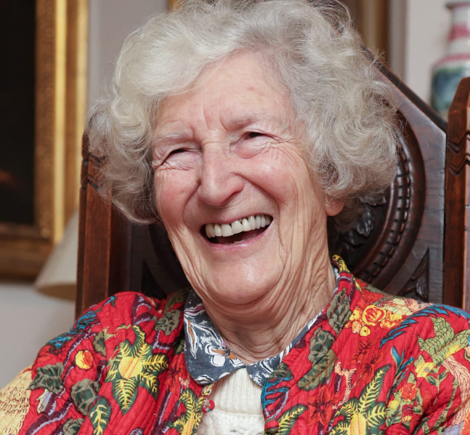 Elderly woman with curly gray hair smiling warmly, wearing a colorful floral jacket, seated indoors. - Home Instead