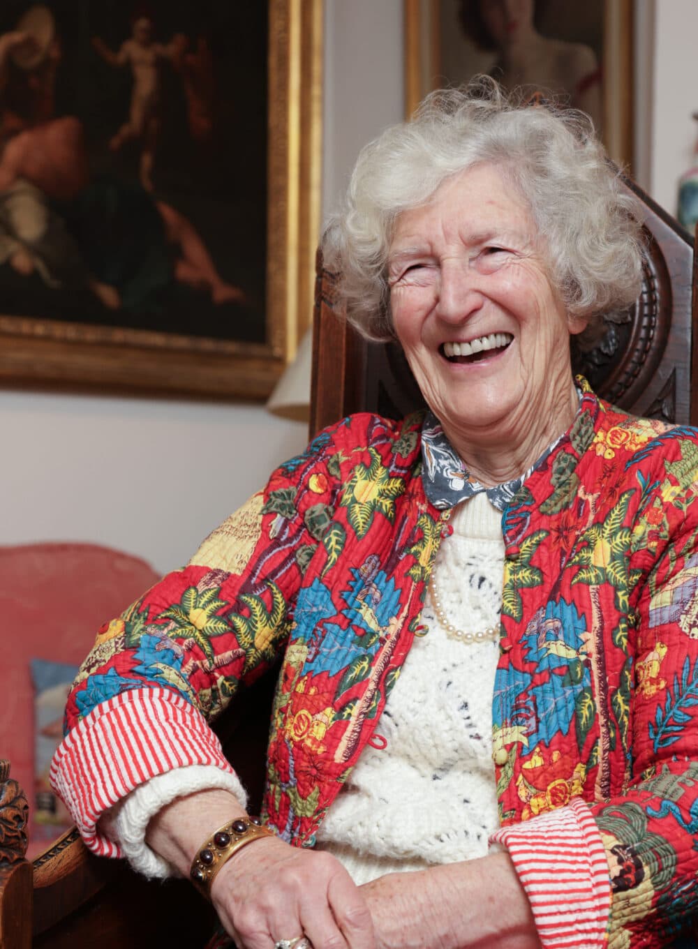 Elderly woman with curly gray hair smiling, wearing a colorful floral jacket, seated indoors. - Home Instead