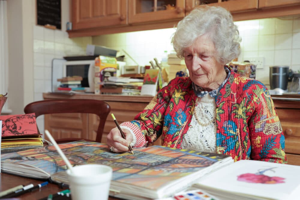Elderly woman painting in a sketchbook at a kitchen table, surrounded by art supplies. - Home Instead
