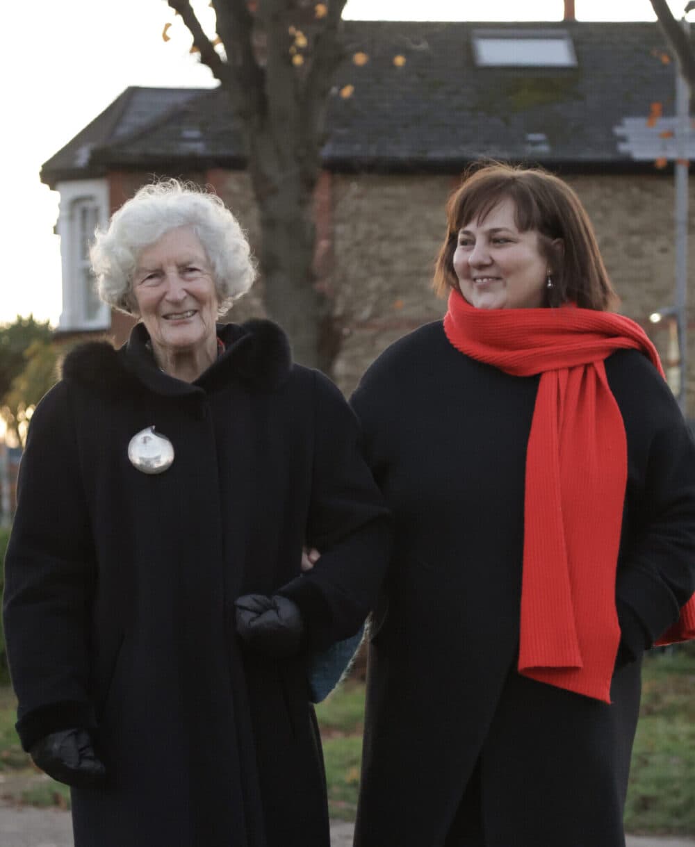 Two women in black coats walking arm in arm outside, one wearing a red scarf, both smiling. - Home Instead