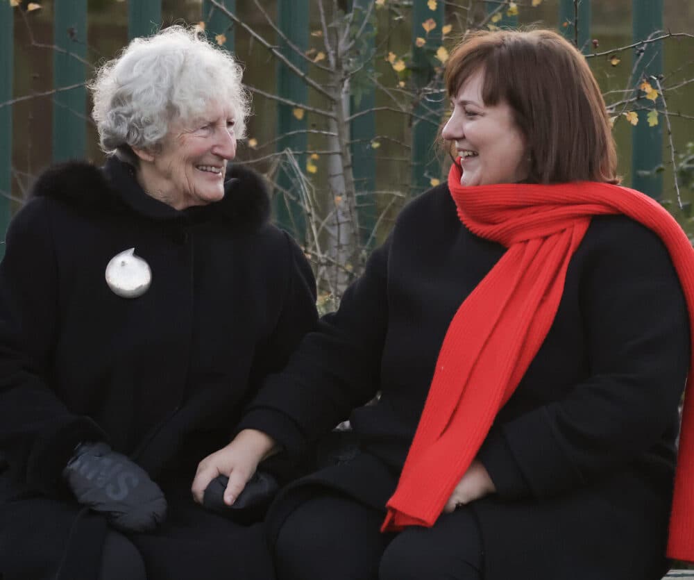 Two women in black coats, one with a red scarf, sit smiling and holding hands outdoors. - Home Instead
