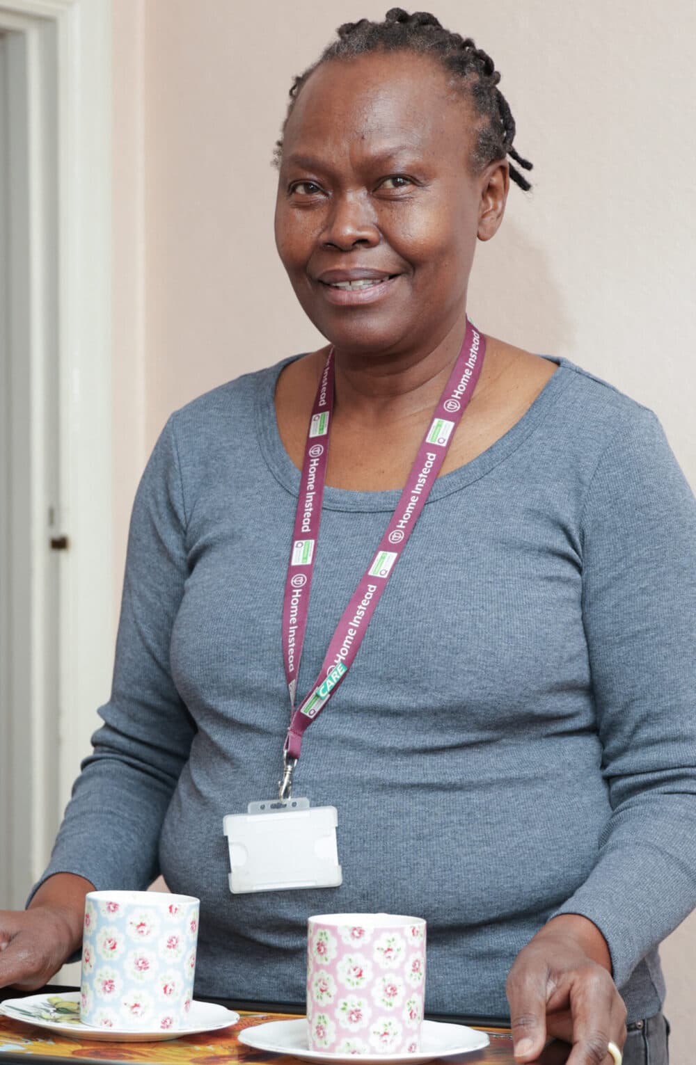 Smiling woman with a lanyard serves two patterned mugs on a tray, standing indoors. - Home Instead