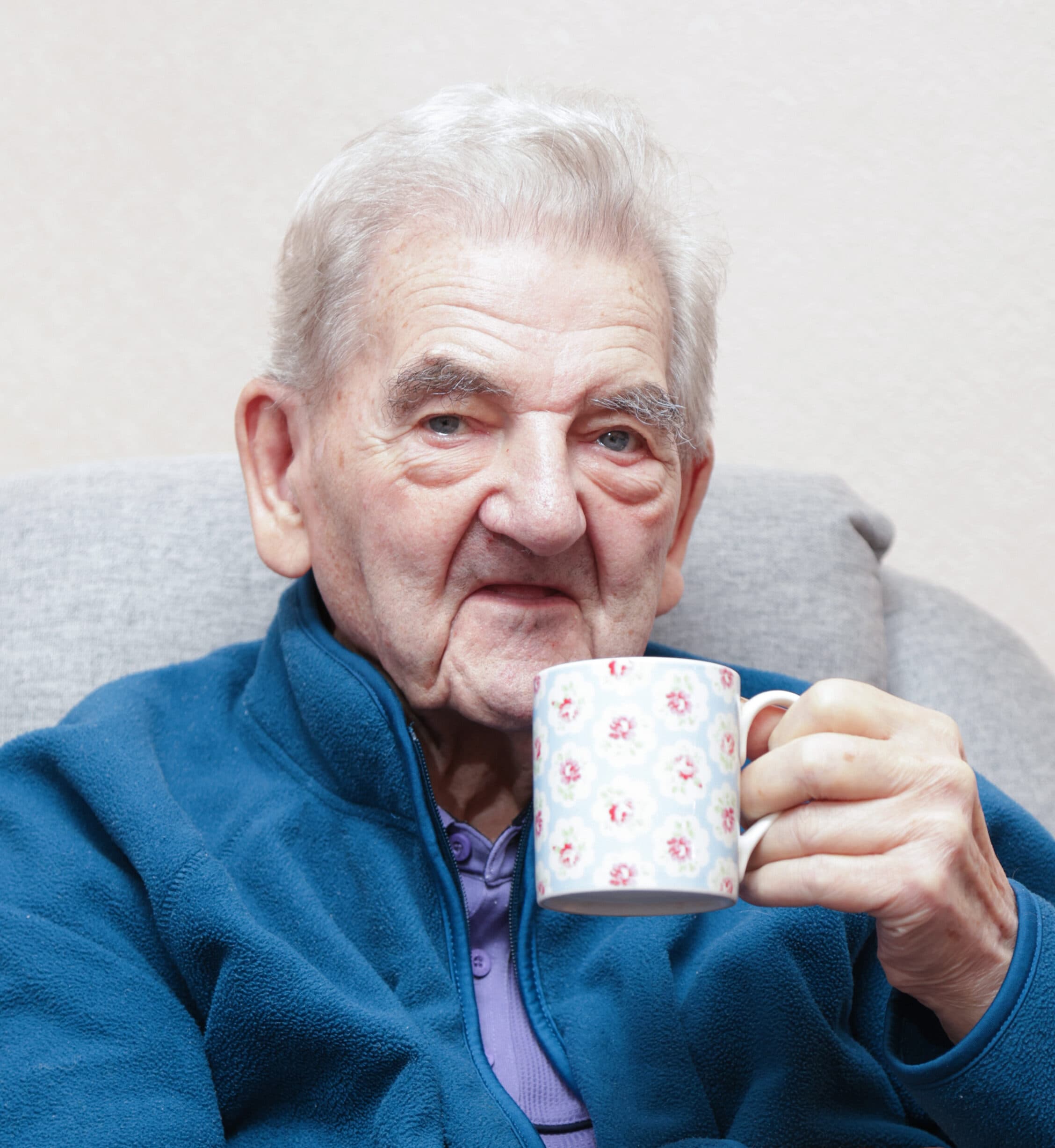 An elderly man in a blue fleece jacket sits on a sofa, smiling and holding a floral mug. - Home Instead