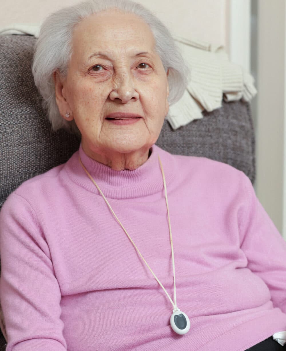 Elderly woman in a pink sweater sitting on a chair, wearing a medical alert pendant around her neck. - Home Instead