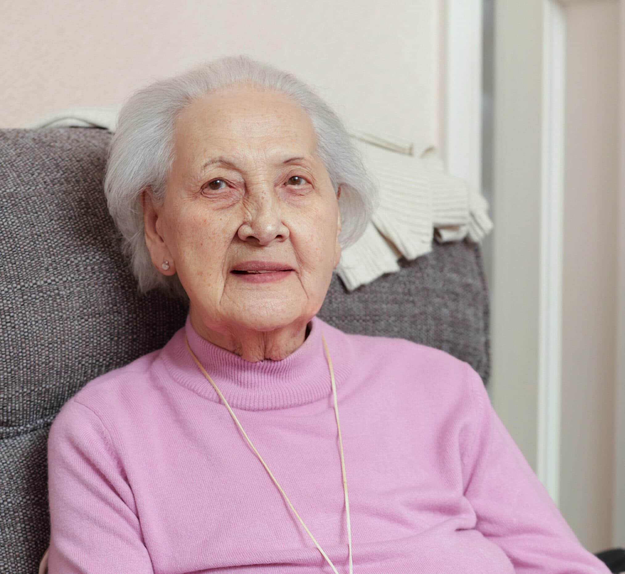 Elderly woman with short gray hair, wearing a pink sweater, sitting on a gray chair and smiling gently. - Home Instead