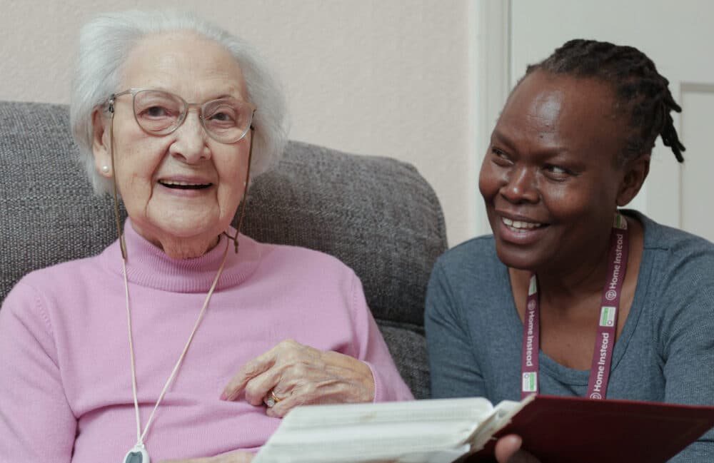 Elderly woman smiling with a caregiver, both sitting and looking at an open book together. - Home Instead