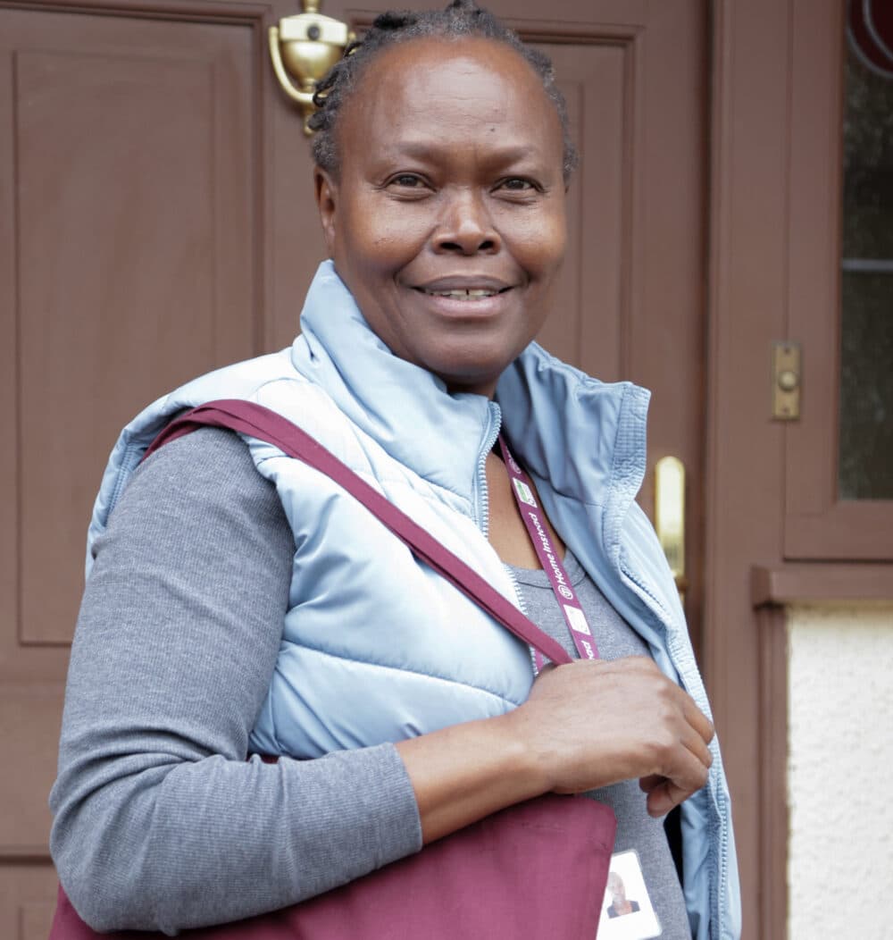 Woman smiling outdoors, wearing a blue vest and gray shirt, holding a maroon bag, standing by a brown door. - Home Instead