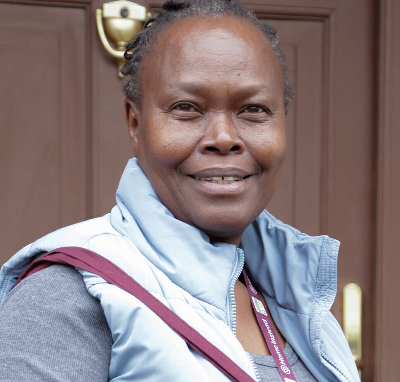 Smiling woman with short hair wearing a blue vest stands in front of a brown door. - Home Instead