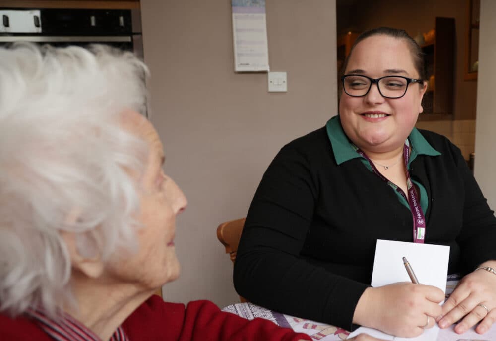 A smiling woman with a notepad talks to an elderly woman with white hair at a table. - Home Instead