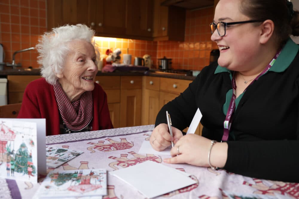 An elderly woman and a caregiver smile and talk at a kitchen table with cards and envelopes. - Home Instead