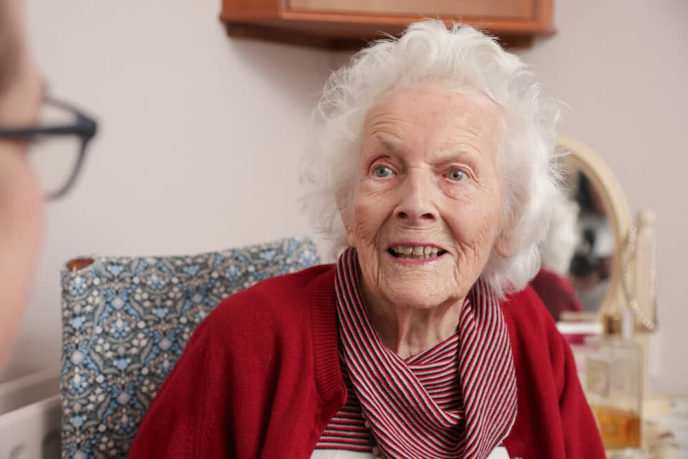 Elderly woman with white hair and red sweater smiling, sitting indoors and talking to someone. - Home Instead